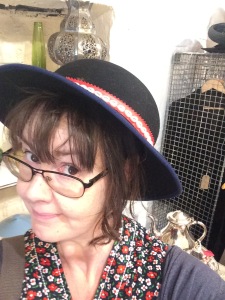Tonia is wearing a vintage hat. She has brown hair with a fringe, black glasses and is half-smiling at the camera. She's also wearing a flowered dress from the 1960s and a blue cardigan. She is in her mid-thirties. 
